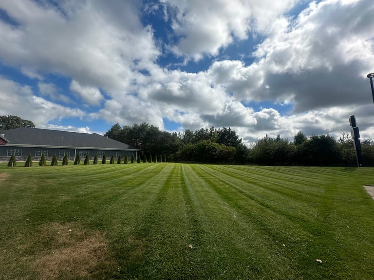Large manicured sports field with diagonal mowing patterns, bordered by a long building and trees under a partly cloudy blue sky